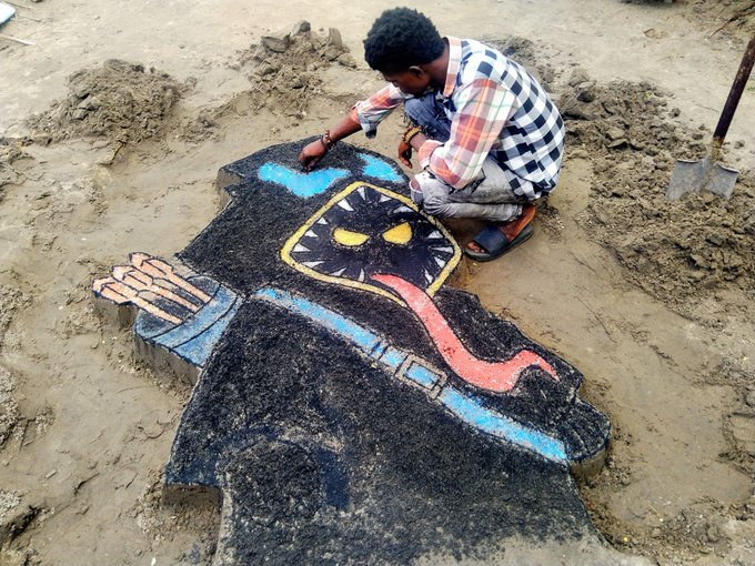 A man kneels on sandy ground, carefully crafting a vibrant sand sculpture of a hooded, cartoonish figure with yellow eyes, a long pink tongue, and outstretched hands, using a shovel visible nearby. The artwork's bold, graffiti-inspired style directly mirrors the post's description of art that resembles the creator's own aesthetic, serving as a tangible tribute to the Hooja NFT collection.