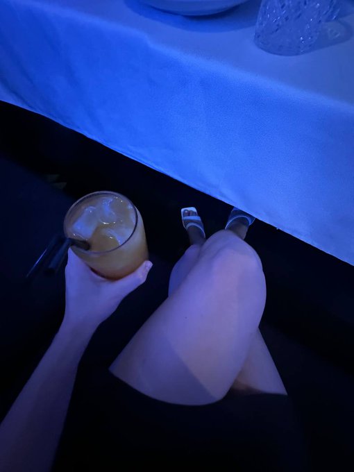 A moody, blue-lit first-person view of a person holding an iced coffee drink while seated with their knees crossed near a white-draped table.