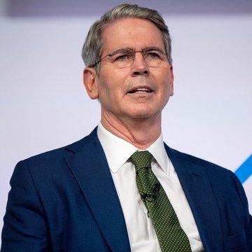 A professional photo of a gray-haired man wearing glasses, a blue suit, and a green patterned tie, speaking at what appears to be a formal event.