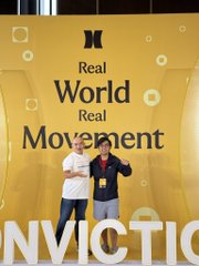 Two people stand smiling in front of a large yellow backdrop featuring the word "CONVICTION" and the text "Real World Real Movement" at a meetup event. The image visually captures the Conviction event mentioned in the post, where the founder of Build Stuffs Saigon and Arc fellowship fellows gathered.