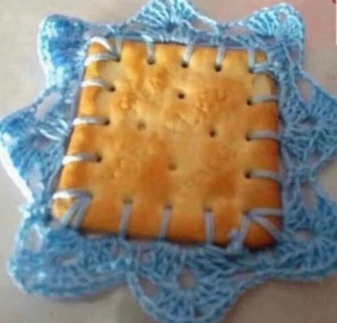 A crocheted textile piece featuring a square cracker-like center with small holes, bordered by light blue scalloped lace edging.