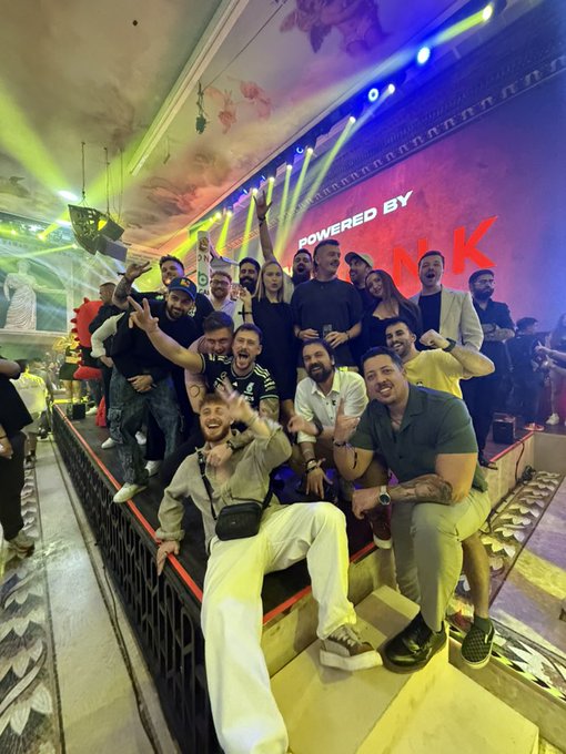 A group of well-dressed friends pose energetically on a staircase at a nightclub event featuring a large illuminated sign reading "POWERED BY NK," with the "GM" post likely referring to their good morning greeting after a night out celebrating together.