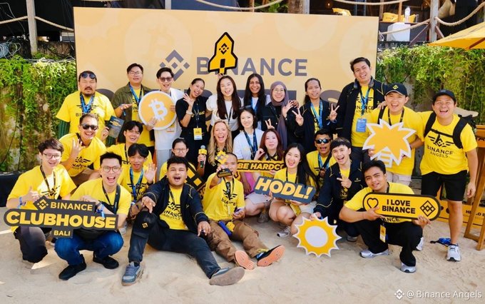 The image shows a group of Binance team members posing together at the Binance Clubhouse event in Bali, with members holding signs displaying "BINANCE CLUBHOUSE #BALI," "#HODL!," "BE HARD!," and "I LOVE BINANCE," all wearing matching yellow Binance-branded shirts in a cheerful, team-celebration setting.