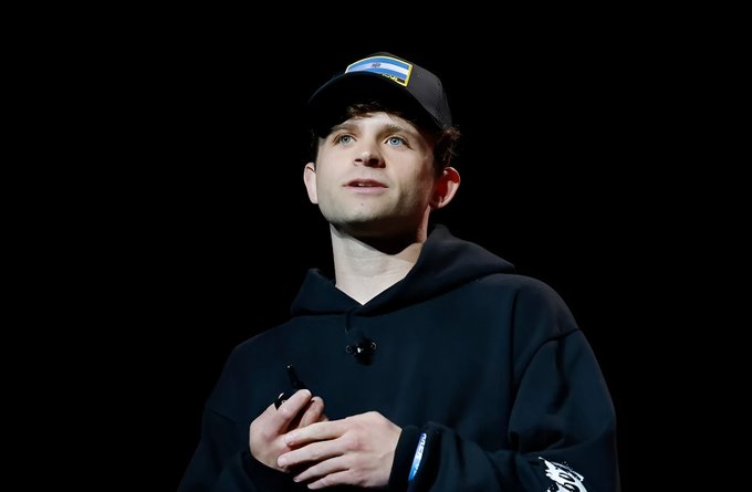 A young man wearing a black hoodie and a black baseball cap with a colorful logo stands on stage against a dark background, looking upward.