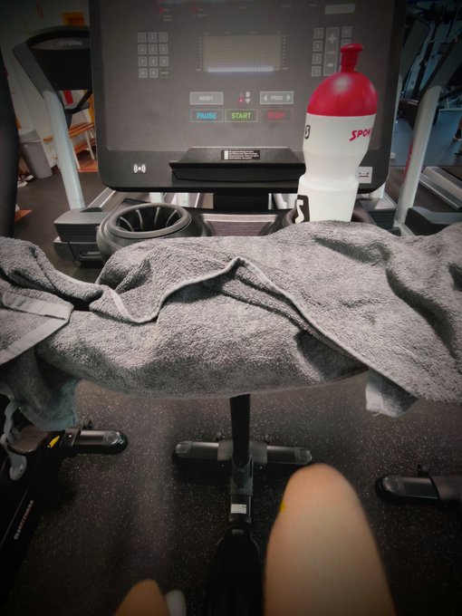 A first-person view from someone sitting on gym equipment, showing a gray towel draped in the foreground with a treadmill and a white water bottle in the background.