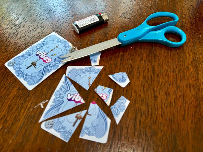 A pair of blue-handled scissors rests on a wooden table, having just cut up a "Vibes" branded playing card and a small battery into multiple pieces. The image humorously illustrates the caption's message "Don't cut my cards moonpay," symbolically showing the destruction of items after the request to block the account.
