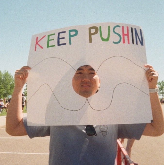 A young man at an outdoor event holds up a handmade sign reading "KEEP PUSHIN" in colorful letters, with a face hole cut between two drawn curves resembling a resistance level on a price chart. The creative sign playfully illustrates the post's caption about waiting for $ZEC to push through resistance past $1000, visually representing a trader's anticipation of a breakout.