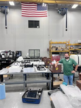 A man in a green shirt stands in an industrial workshop next to engine parts, with an American flag hanging on the wall behind him.