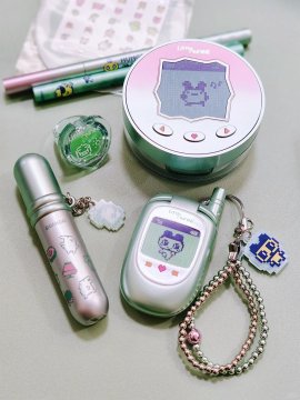 A flat-lay arrangement of cute stationery and accessories including a pink Tamagotchi-style device, green pens, lip gloss, and character charms.