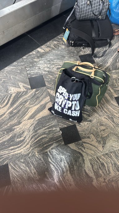 A green duffel bag with a black tote reading 'SECURE YOUR CRYPTO LIKE CASH' sits on a polished marble floor in what appears to be an airport or transit area, with luggage visible in the background.