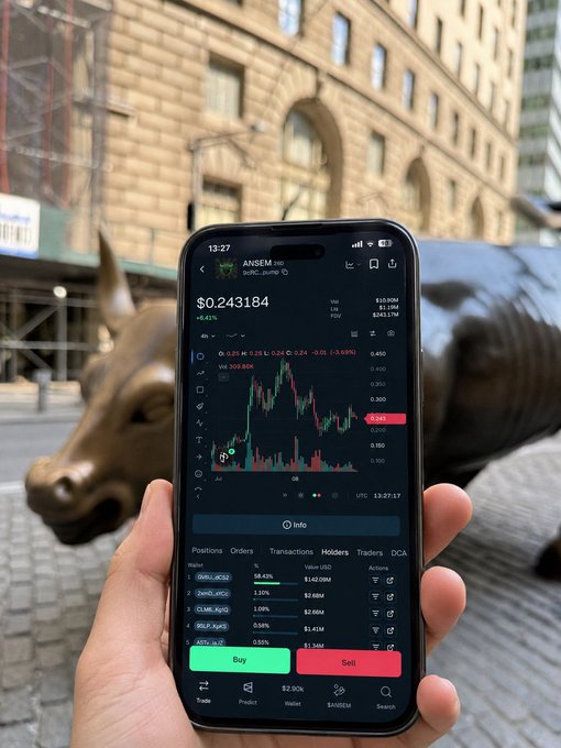 A hand holds a smartphone displaying a cryptocurrency trading chart for 'ANSEM' at $0.243184 in front of the Wall Street Bull statue.