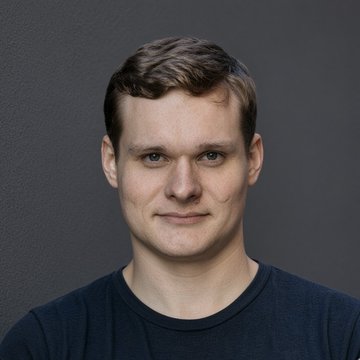 Professional headshot portrait of a smiling man with short brown hair wearing a dark navy t-shirt against a gray background.