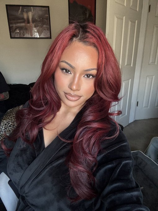 A woman with long, wavy burgundy hair poses for a selfie in a dark robe, with a framed picture visible on the wall behind her.