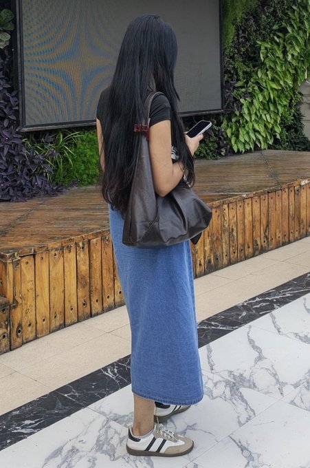 A woman with long black hair stands on a marble floor holding a phone and a brown bag, wearing a black shirt, long denim skirt, and Adidas Samba sneakers.