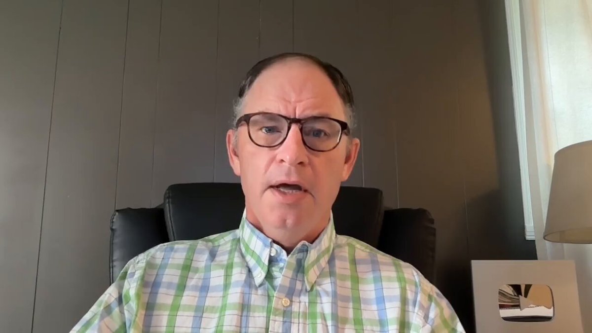 The image shows a man with short hair wearing glasses, sitting in a chair at a desk or table. He has his mouth open and appears to be giving an explanation about the Bitcoin price. The backdrop for this scene is a gray wall that complements the color of the man's shirt. In addition to the man, there are two more people in the frame: one standing near him on the left side, and another person sitting further away at the top right corner of the image.