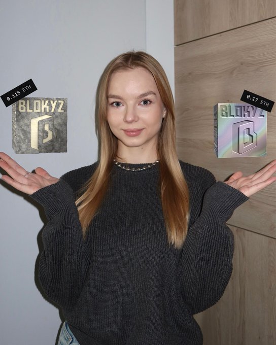 A young woman with long blonde hair holds up two NFT packages labeled "BLOKYZ" with prices of 0.115 ETH and 0.17 ETH displayed above them, celebrating her recent purchase from the Blokyz project. She wears a dark gray sweater and a beaded necklace, with her hands outstretched in a presenting gesture as she poses in front of a wood-paneled wall.