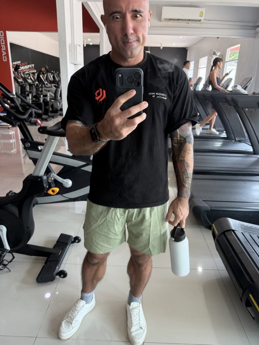 A bald, tattooed man wearing a black t-shirt and green shorts takes a mirror selfie with his phone while holding a light blue water bottle in a gym setting.