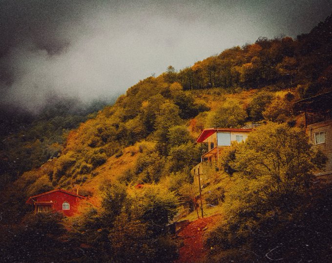 A misty hillside landscape shows autumn-colored trees with small red-roofed houses nestled into the mountainside under a cloudy sky.