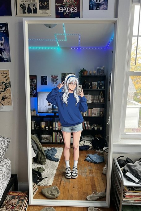 A person wearing a blue sweatshirt, denim shorts, and a white-haired anime cosplay wig with headphones takes a mirror selfie in a bedroom with LED lights and Hades posters on the wall.