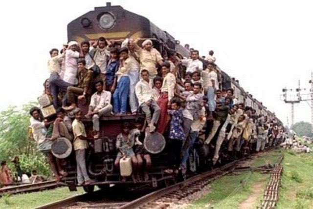 A heavily overcrowded train is packed with people riding on top of and hanging from the sides of the train cars on the tracks.