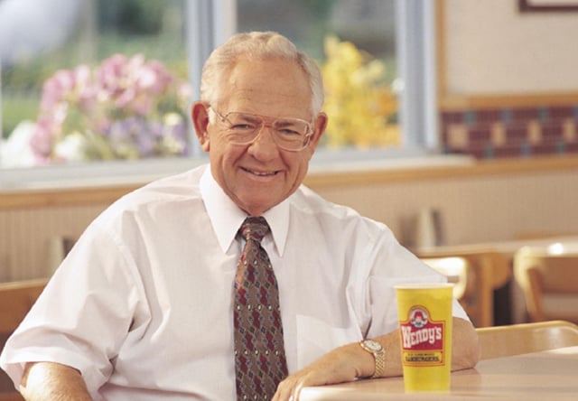 An elderly man in a white shirt and patterned tie smiles at a Wendy's restaurant table, with a yellow Wendy's cup labeled "Wendy's" and "The Official Hamburger" beside him. The image pays tribute to Dave Thomas, the founder of Wendy's (ticker: WEN), who is affectionately known as "Big Dave," connecting to the post about purchasing shares in his memory.