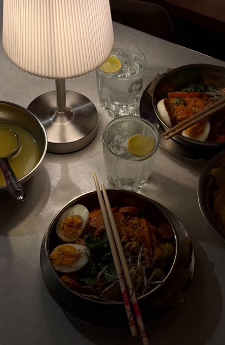 A dimly lit table setting with two bowls of noodles topped with halved boiled eggs and vegetables, served with chopsticks, alongside glasses of water with lemon slices and a small lamp.
