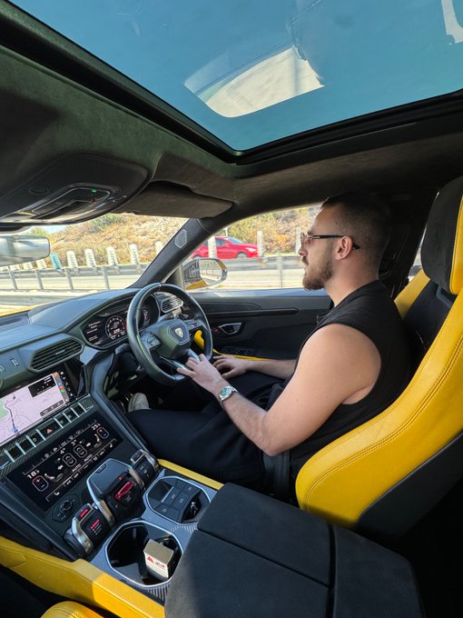 A man sits in the driver's seat of a yellow Lamborghini Urus, holding the steering wheel while the panoramic sunroof reveals a bright blue sky overhead, with a red vehicle parked nearby in the background.
