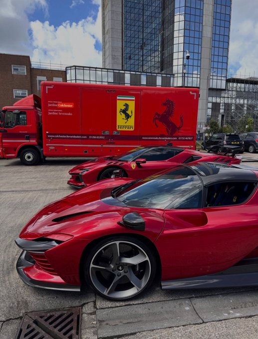 Two glossy red Ferrari sports cars are parked in front of a matching red Ferrari transport truck displaying "Jardine Colchester," the iconic Ferrari prancing horse logo, and contact details for "www.carsalesferrari.co.uk." The image showcases Ferrari's signature red branding and luxury vehicles, aligning with the post's appreciation shared via the Pumpfun app.