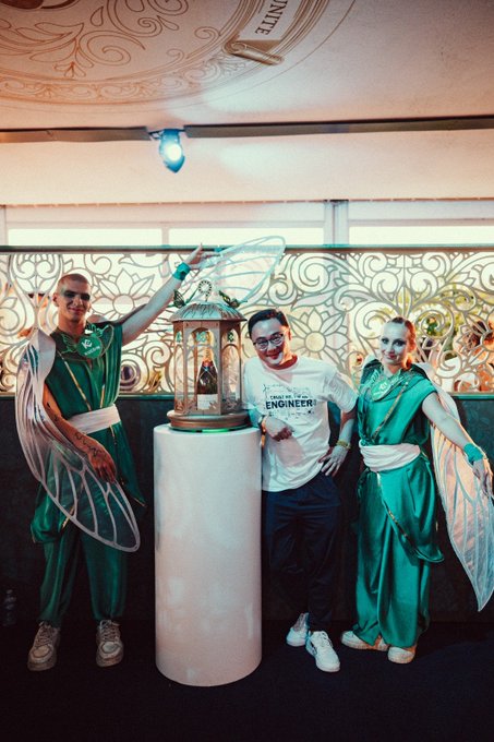 A man wearing a "TRUST ME I'M AN ENGINEER!" t-shirt poses between two performers dressed as fairies in green and white costumes, with an ornate lantern on a glowing pedestal, capturing a magical moment from the Tomorrowland festival's On Cloud 9 Skybox Experience anniversary celebration.