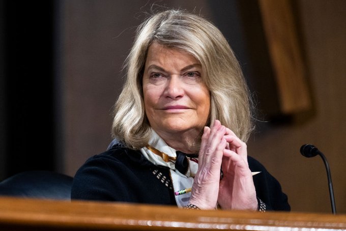 An older woman with shoulder-length blonde hair sits at a desk with a microphone, hands clasped together in front of her.