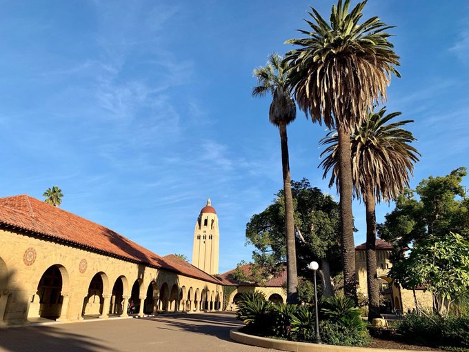 A sunlit view of a Spanish mission-style campus with arched colonnades, a red-tile roof, and a tall central tower, framed by tall palm trees against a blue sky.