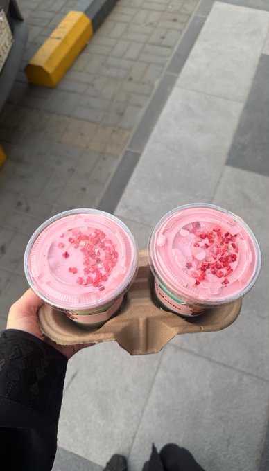A first-person view of a hand holding a cardboard tray with two plastic cups containing pink beverages topped with red crumbles, likely strawberry-flavored drinks, against a background of a paved sidewalk.