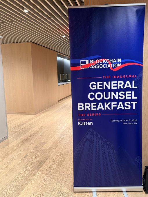A blue vertical banner for the 'BLOCKCHAIN ASSOCIATION THE INAUGURAL GENERAL COUNSEL BREAKFAST THE SERIES,' sponsored by Katten, taking place Tuesday, October 6, 2026 in New York, NY, displayed in a modern interior space with wood flooring.