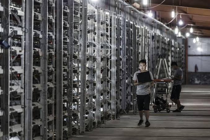 A young man walks down an aisle in a server farm/data facility carrying a laptop, with rows of computer servers lining both sides.