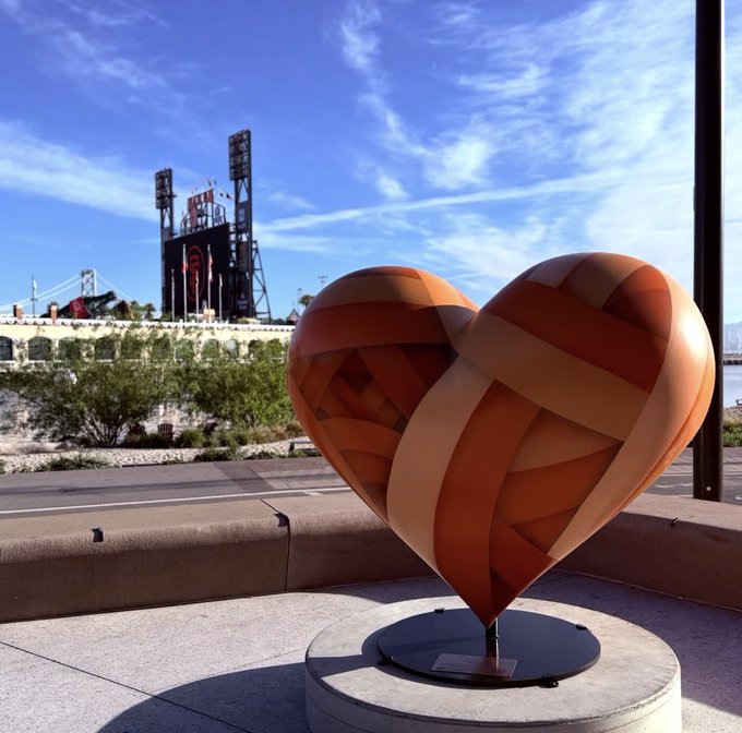 A heart-shaped sculpture with orange and tan swirling stripes sits on a pedestal, with the Oracle Park stadium and its large scoreboard visible in the background under a blue sky.