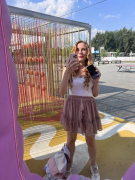 A woman takes a mirror selfie while holding a phone, wearing a white tank top and pink skirt in an outdoor setting with colorful hanging decorations behind her.