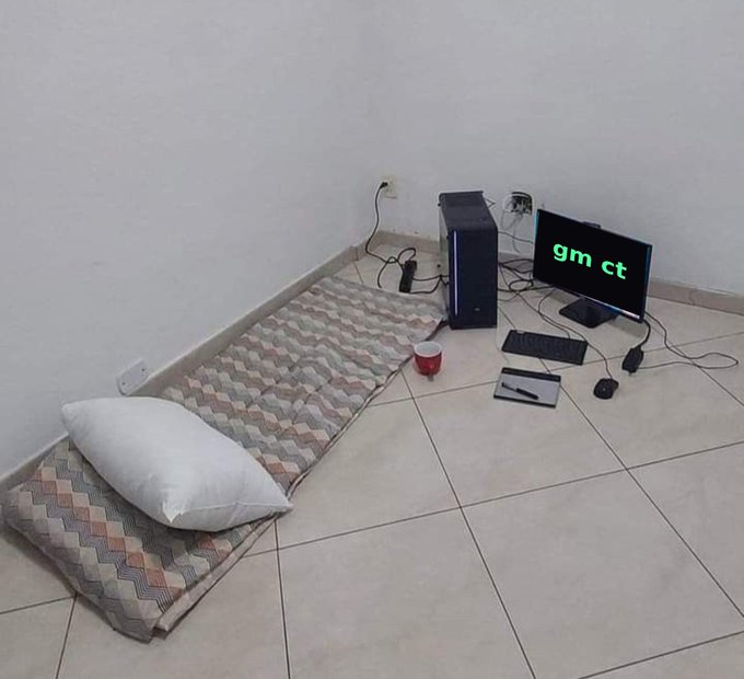 A minimalist floor setup features a thin mattress, pillow, and red cup alongside a desktop monitor displaying 'gm ct' in green text.