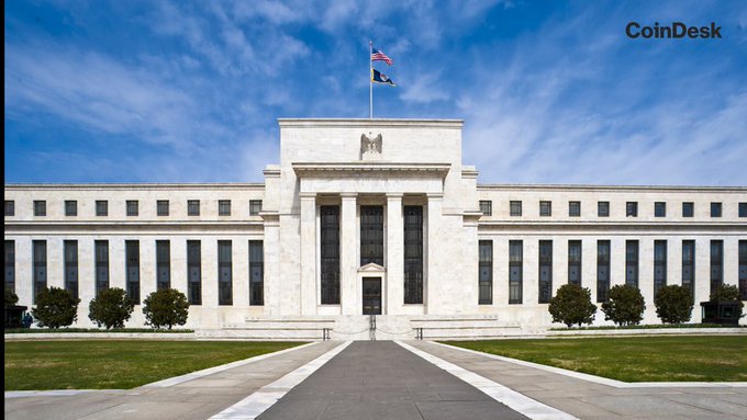 The image shows the Federal Reserve building in Washington D.C. with American flags flying overhead, watermarked with "CoinDesk" in the upper right corner. The imposing neoclassical architecture of the central bank symbolizes its monetary policy authority, directly relevant to the post's discussion of U.S. jobless claims coming in below forecast at 199K and the implications for the Fed's interest rate decisions ahead of the payrolls report.