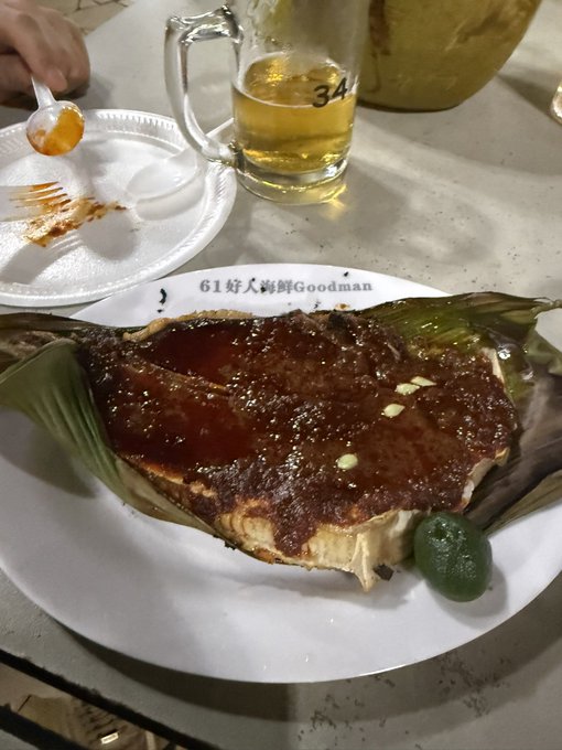 A piece of glazed meat served on a banana leaf atop a white plate labeled '61 好人海鲜Goodman', accompanied by a glass of beer and a partially eaten styrofoam plate.