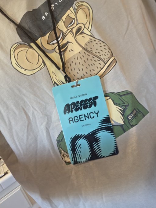 A blue event badge reading “APERTUR AGENCY” hangs on a lanyard over a shirt featuring a large illustrated bear-like character.