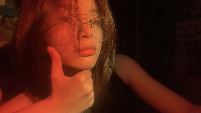A dimly lit selfie of a young woman wearing glasses giving a thumbs-up gesture, with red lighting illuminating her face.