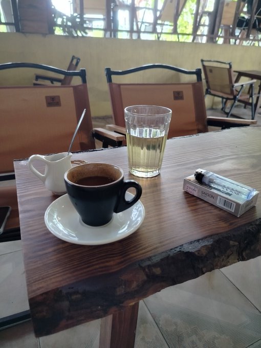 A small black cup of Vietnamese coffee sits on a saucer next to a glass of tea and a small white creamer pitcher on a wooden table, with a pack of Saigon cigarettes resting nearby.