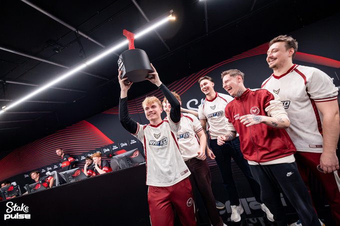 An esports team in white and red jerseys celebrates by hoisting a trophy, with a 'Stake pulse' watermark in the bottom corner.