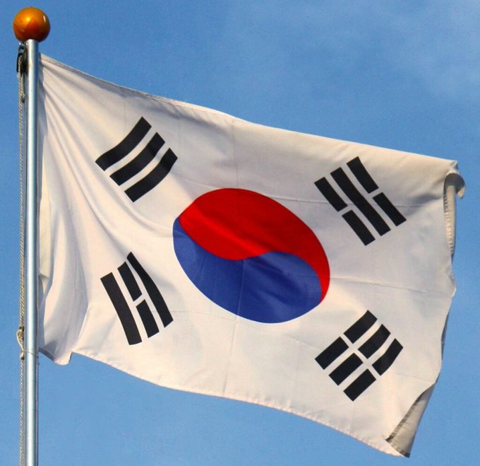 The South Korean national flag, known as the Taegeukgi, waves against a clear blue sky, featuring the red and blue Taegeuk symbol in the center and four black trigrams in the corners.