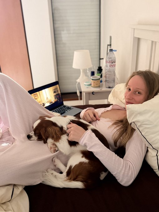 A woman lies on a bed petting a brown and white Cavalier King Charles Spaniel, with a laptop displaying a video and Evian water bottles on a nightstand nearby.