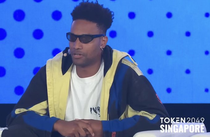 A young man wearing sunglasses and a colorful yellow, blue, and black jacket speaks on stage at TOKEN 2049 Singapore, with a blue polka-dot backdrop behind him. The image relates to a post highlighting his recent earnings as a founder, listing $187,332 in revenue across Bullpen and Ansem Launchpad over the past 30 days.