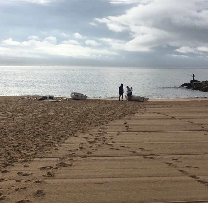 The image shows a beach scene with people standing near boats and water. There are two boats visible - one closer to the left side of the frame and another further back, towards the right side. The sky above them appears cloudy, suggesting that they might be at the beach during a slightly overcast day. A person is also present in the image, possibly observing or preparing for boating activities.