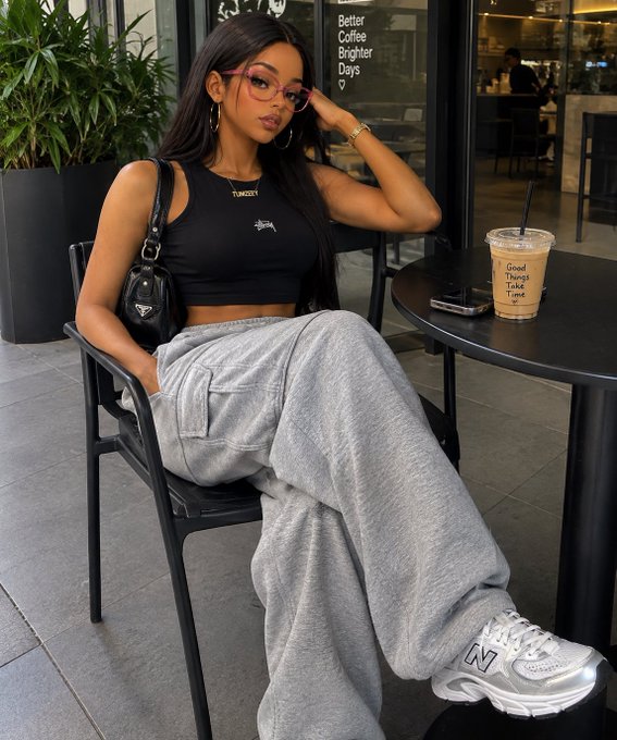 A woman wearing a black Stüssy crop top, grey sweatpants, and New Balance sneakers sits at an outdoor cafe table with a 'Good Vibes Take Time' iced coffee cup beside her.
