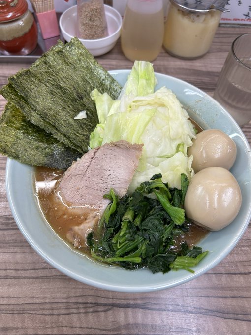 A bowl of ramen with nori sheets, chashu pork, cabbage, spinach, and two marinated eggs in a light broth, with condiment jars and a glass of water in the background.