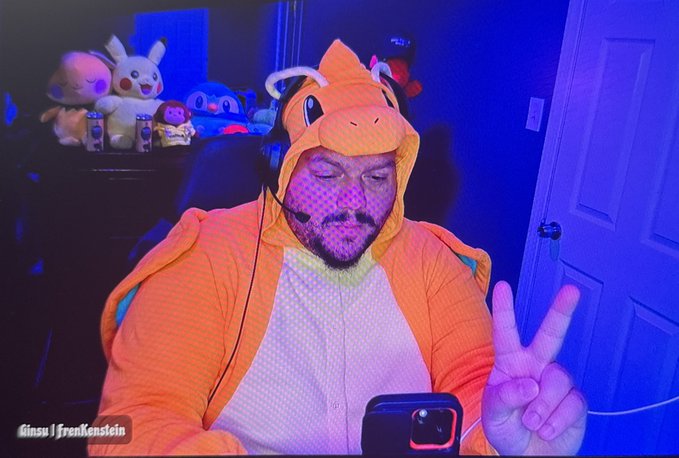 A man wearing an orange Charizard onesie costume and headphones, making a peace sign while on a video call, with plush toys visible on a shelf behind him in blue-lit room; the image is watermarked "Ginsu | Frenkenstein."